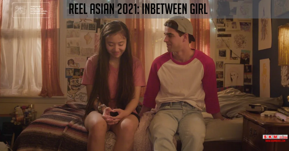 A still from InBetween Girl. A mixed Asian American teen girl sits next to a Caucasian American teen boy on a bed in the teen girl’s bedroom. He’s looking at her. She’s looking down. Both are smiling.